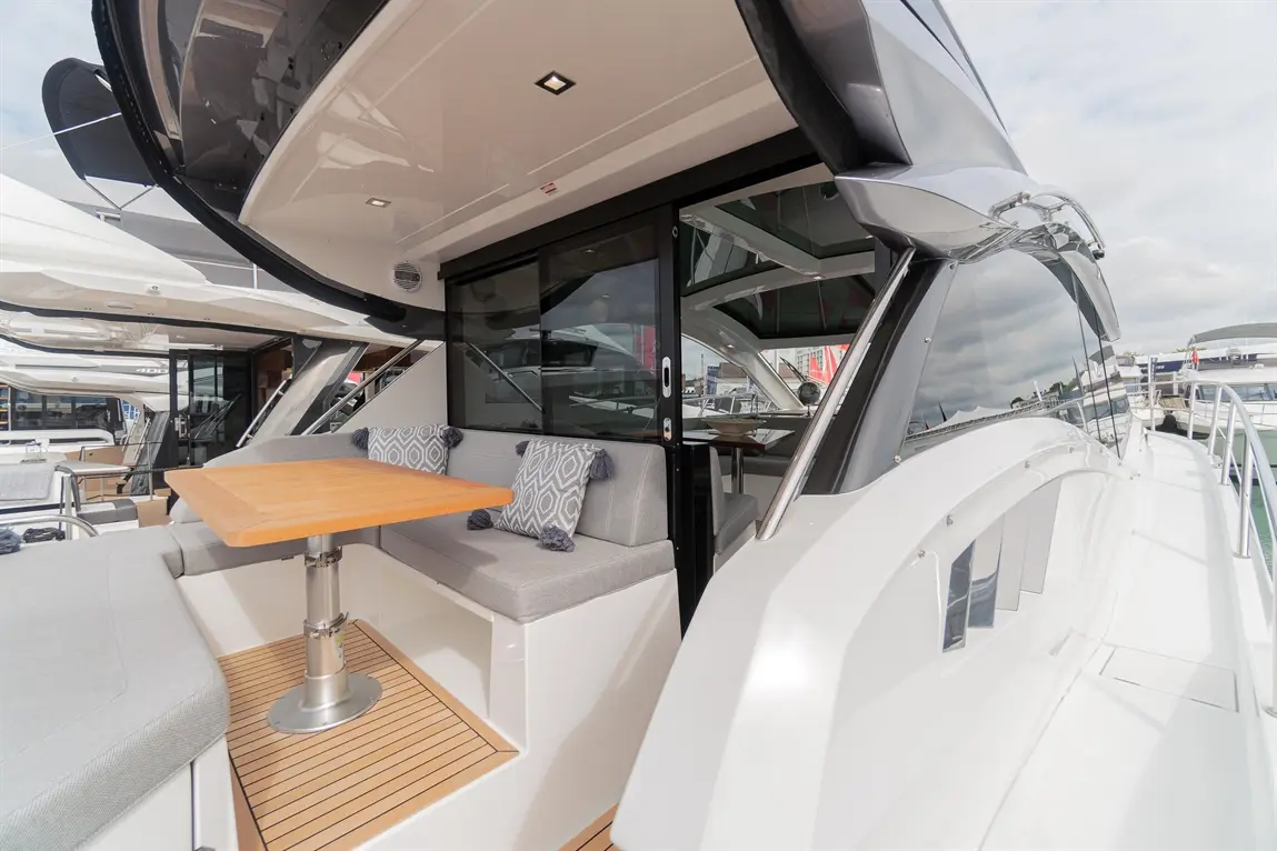 Modern yacht deck with a wooden table, gray upholstered seating, and large windows overlooking the marina and water.